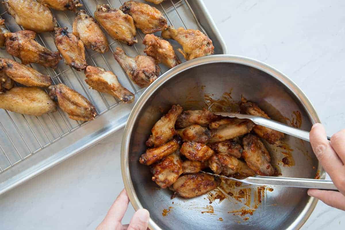 The poultry is tossed in the sauce in a metal bowl with a pair of tongs. 