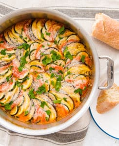 An overhead view of a pan filled with the baked Italian Ratatouille. A piece of bread sits on a white plate to the bottom right. A broken loaf of bread peeks out from the top right hand side of the image.