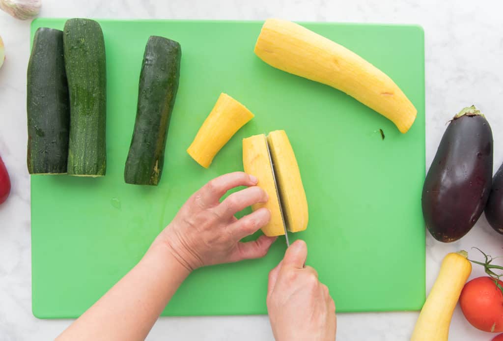 A hand holds a yellow summer squash while another hand holds a knife used to slice it in half. Zucchini sits at top left eggplants and tomatoes are at bottom right