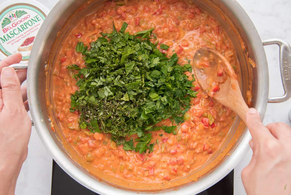 A large pile of green herbs on the tomato-mascarpone sauce in a round pan. A hand uses a wooden spoon to stir the sauce while the other hand holds the pan. A white container of Belgioioso mascarpone is at top left