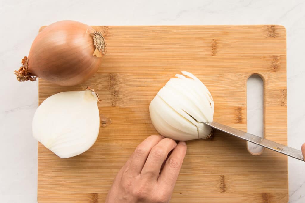 Two hands thinly slice yellow onions on a wood cutting board.