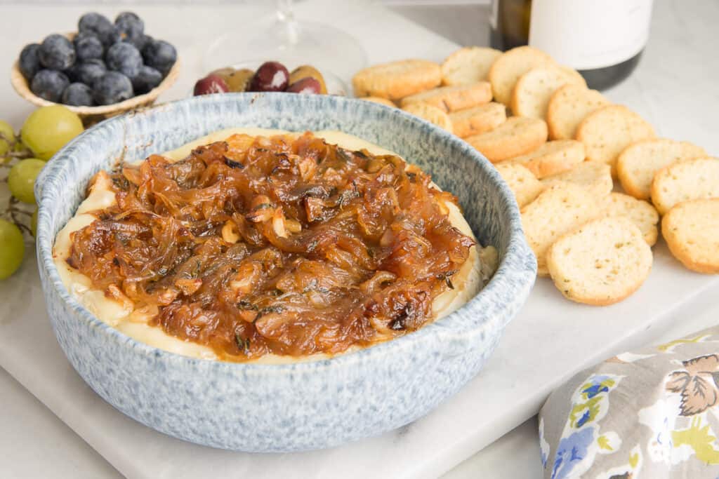 Horizontal image of a blue speckled bowl of baked brie topped with caramelized onions next to grapes, blueberries, and baguette chips