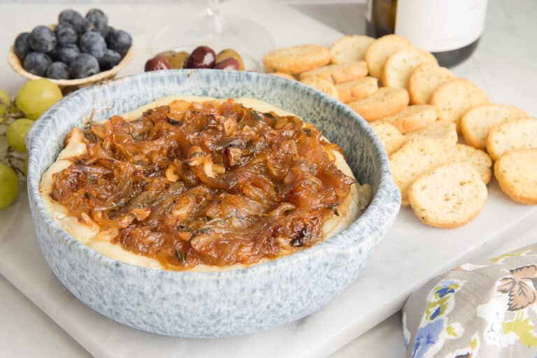 A bowl of baked cheese topped with caramelized onions sits on a marble surface. Surrounding it are slices of toasted bread, grapes, figs, and blueberries, creating an inviting appetizer spread.