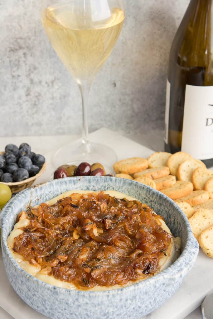 Baked brie in a blue baking dish next to a glass of white wines and accoutrements