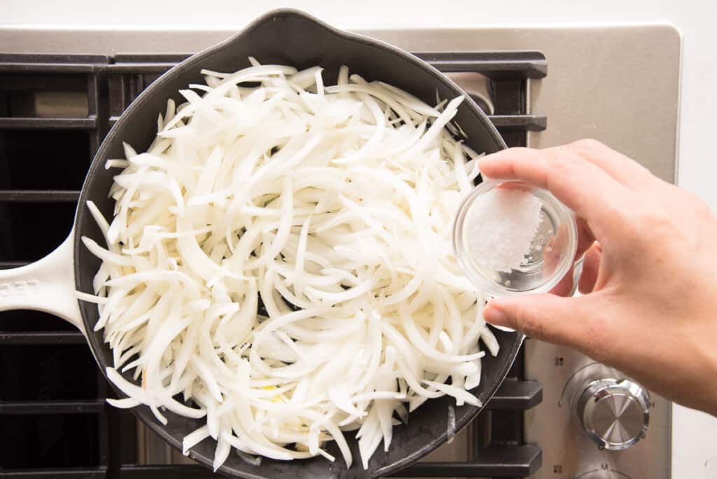 A hand adds salt to the sauté pan of onion slices. 