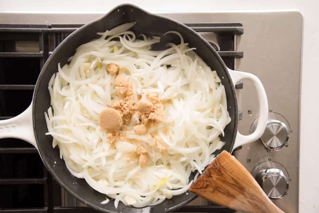 Brown sugar added to a black sauté pan of white onion slices. 