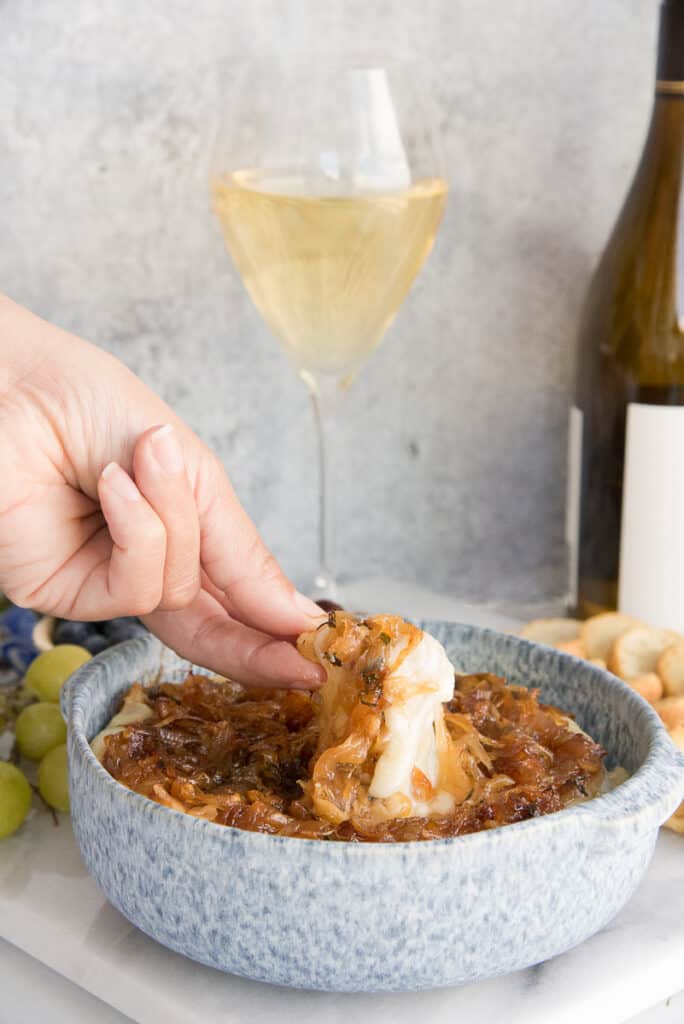 Lead image of a hand using a baguette chip to scoop up the Baked Brie Topped with Caramelized Onions.