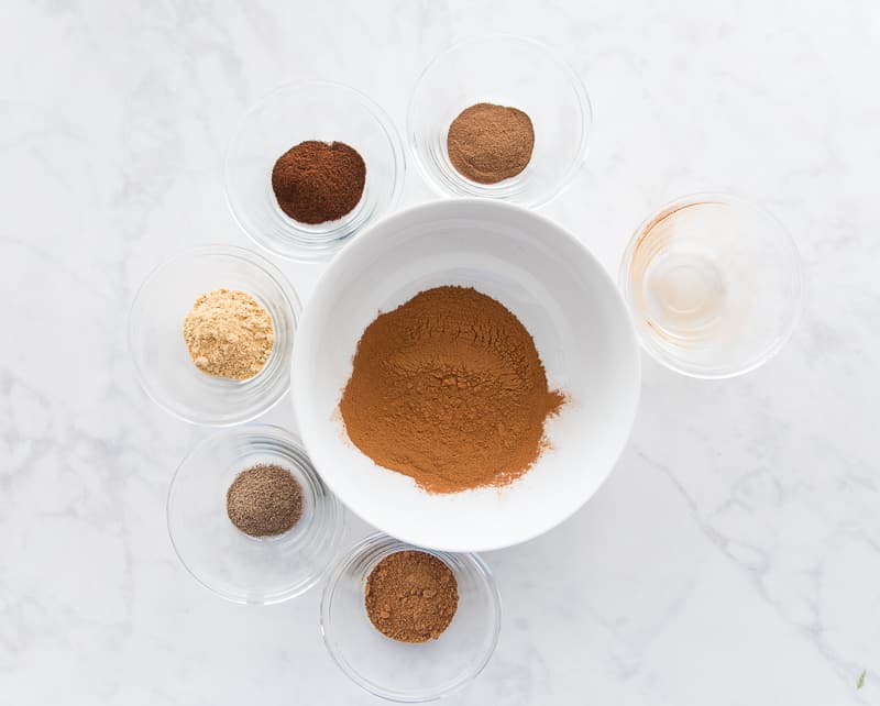 Cinnamon is added to a white mixing bowl which is surrounded by the rest of the spices.