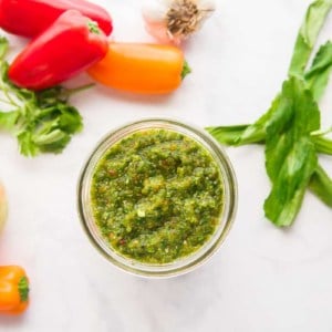 A horizontal image of a jar of Sofrito surrounded by the ingredients used to make it.