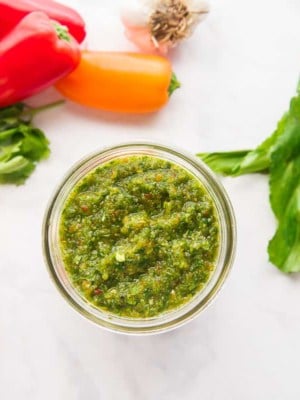 A horizontal image of a jar of Sofrito surrounded by the ingredients used to make it.