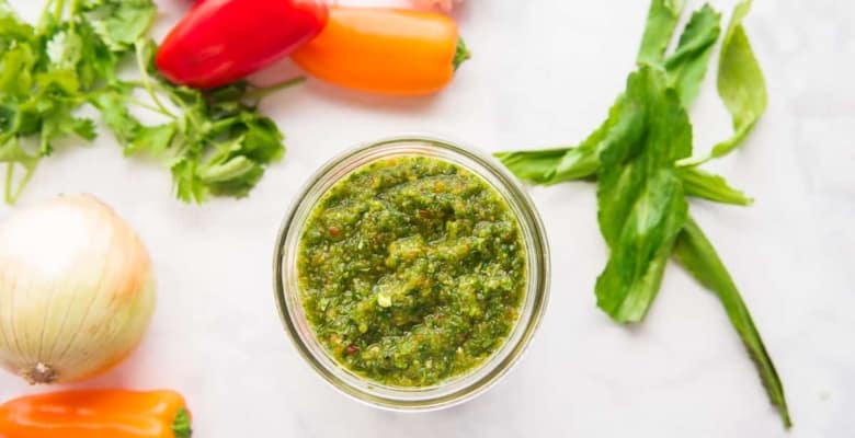 A horizontal image of a jar of Sofrito surrounded by the ingredients used to make it.