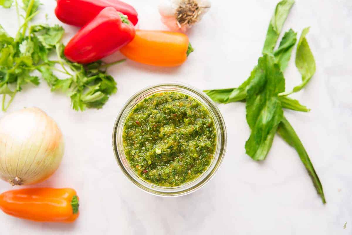 A horizontal image of a jar of Sofrito surrounded by the ingredients used to make it.