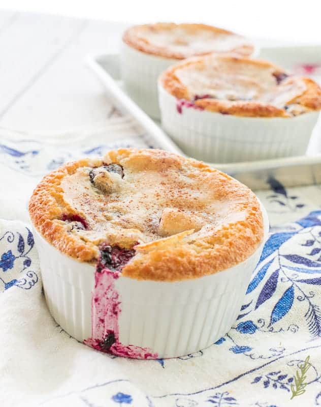 A Blackberry Cobbler in a white ramekin has a drizzle of blackberry juiced baked to the outside of the white dish. The remaining cobblers are on a sheet pan in the top right corner. 