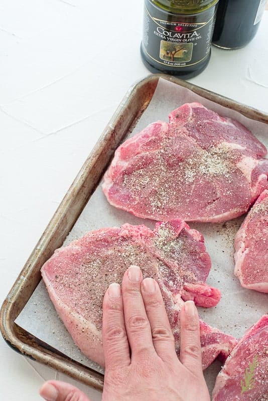 A hand rubs the spice mixture into the pork chops which are on a white piece of paper on a sheet pan. The bottle of olive oil is top center