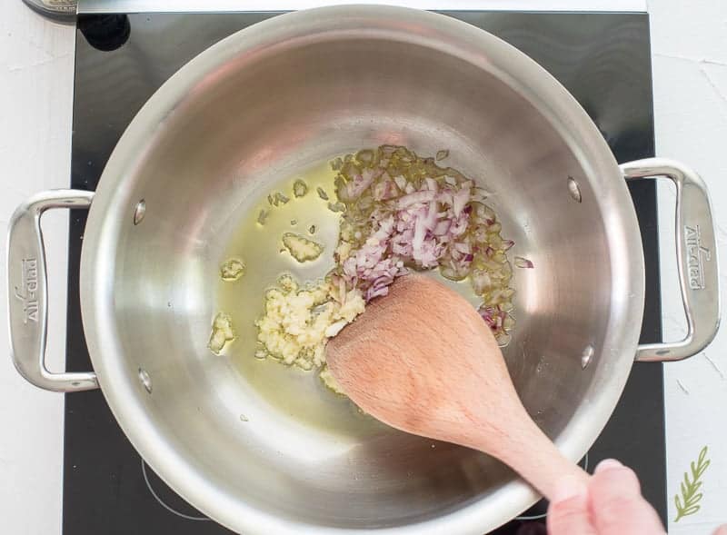 A hand stirs the onion and garlic into hot oil in pot using a wooden spoon.