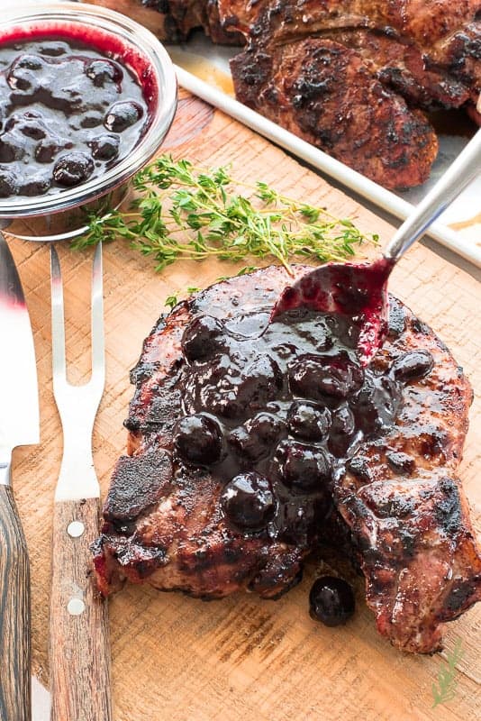 A spoon ladles more Blueberry Balsamic BBQ Sauce onto an individual pork chop which is on a wooden board. 
