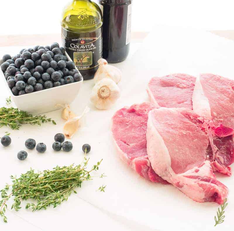 Ingredients shot: stems of fresh thyme, a white bowl of blueberries, fresh garlic, olive oil, balsamic vinegar, and double-cut pork chops on white butcher's paper. 