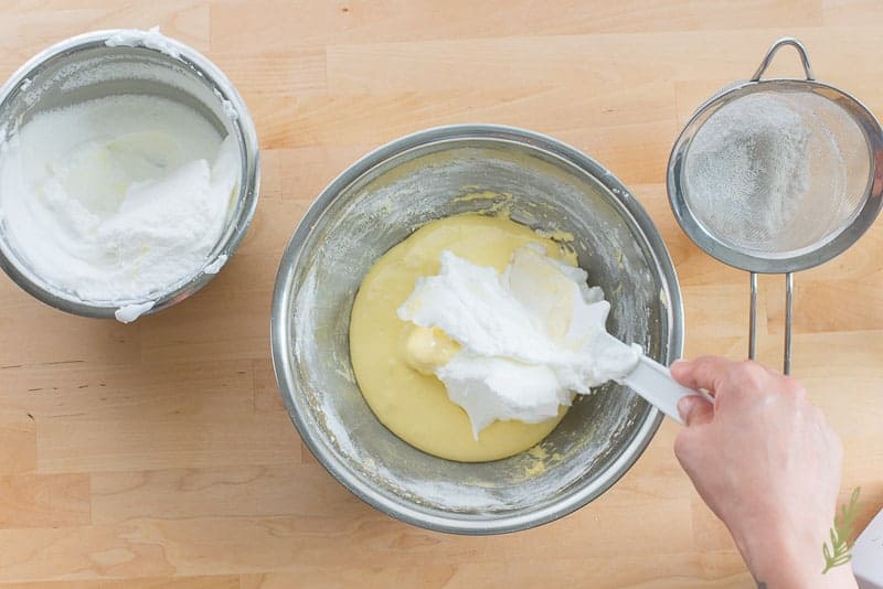 A hand uses a white spatula to fold whipped egg whites into the flour-egg yolks mixture in a metal bowl. 