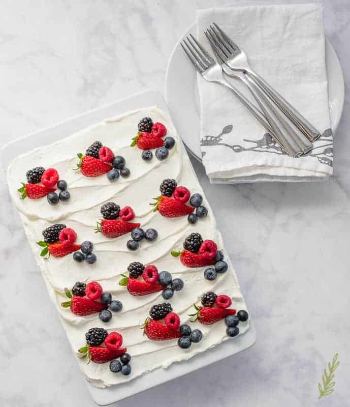 A white baking dish is filled with Berry Tiramisu. It is topped with berries. Next to white plates with napkin and forks on it. 