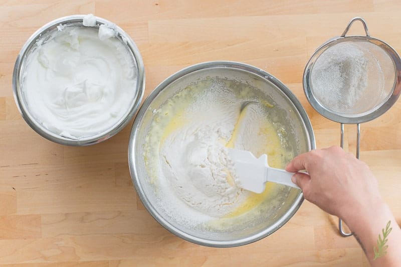 A hand uses a white spatula to fold flour mixture into egg yolks.
