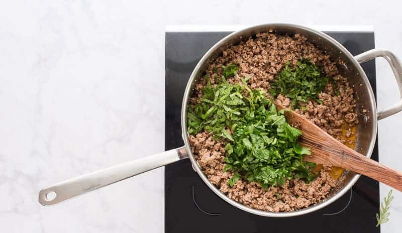 The fresh herbs are added to the meat in the pan. A wooden spoon is used to stir them in