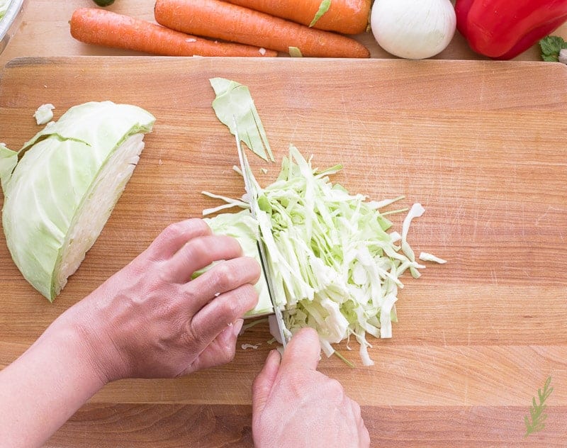 A hand finely shreds green cabbage for Peruvian Inspired Coleslaw with a knife on a wooden cutting board. 
