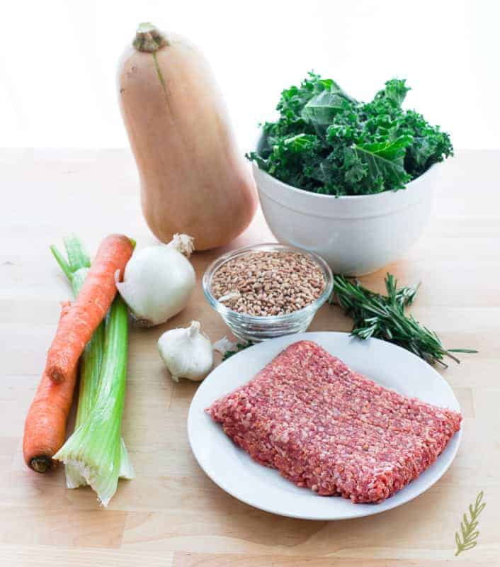 Butternut squash, a white bowl of kale, a white plate of ground sausage, herbs, carrots, celery, onion, garlic and a glass bowl of farro on a wooden surface