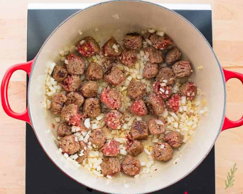 Sausage meatballs, onion, and garlic are sauteed in a red dutch oven. 