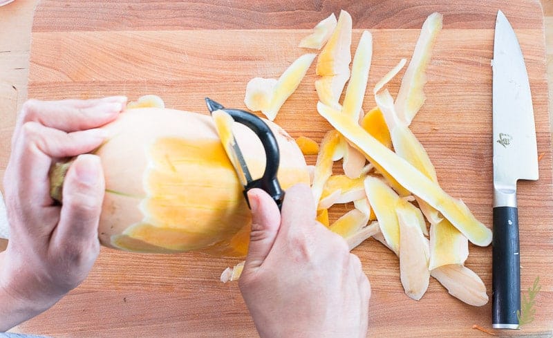 A hand peels a butternut squash with a vegetable peeler on a wooden cutting board. 