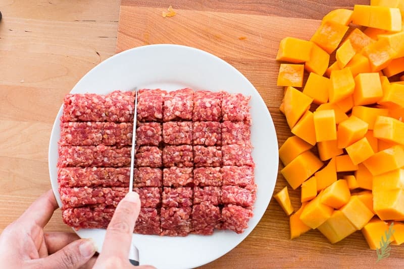 A hand uses a knife to cut ground sausage into chunks on a white plate.