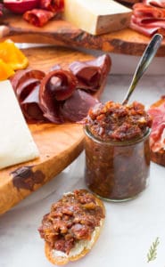 A side view of a small jar of Bacon Bourbon Jam with charcuterie in the background