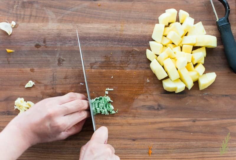 A hand uses a knife to chiffonade sage leaves on a dark wooden surface a pile of diced apples is to the right.