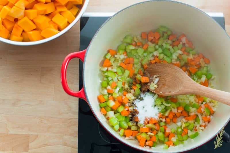 Diced carrots, celery, and onions are sauteed in butter and oil in a red dutch oven. A wooden spoon stirs in salt and pepper. 