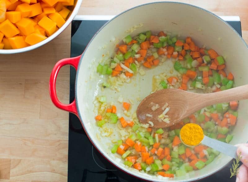 A measuring spoon of curry powder is being added to the dutch oven of sauteed carrots, celery and onion.
