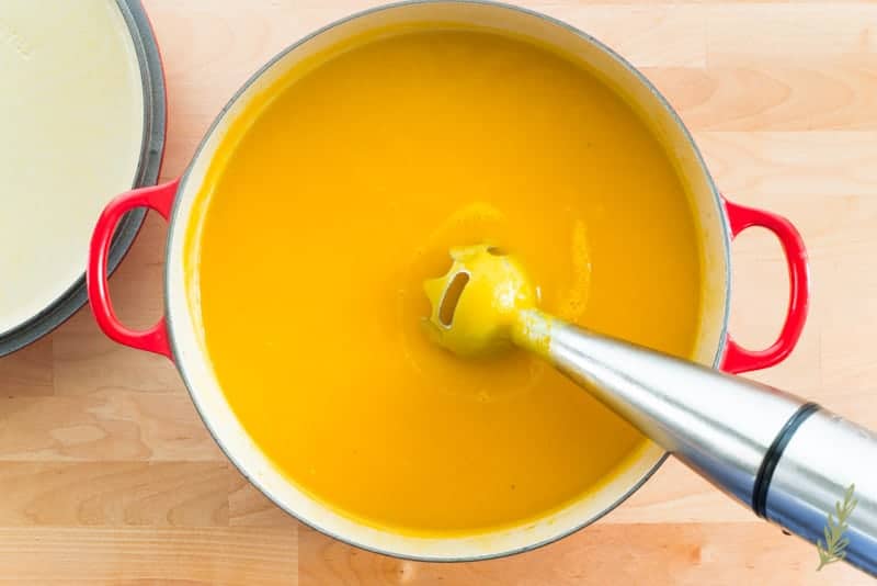 An immersion blender is coming out of the Butternut-Apple Bisque after having pureed it. 