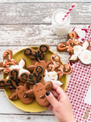 A hand holds a Gingerbread Reindeer above a yellow plate filled with more cookies. A glass jar of milk has a red and white straw in it.