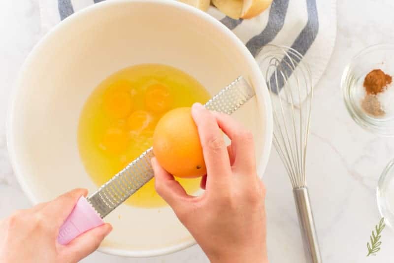 A hand zests the peel of an orange into eggs using a silver microplane