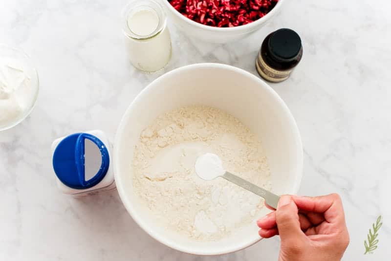 A hand measures baking powder into a white mixing bowl filled with the rest of the dry ingredients. 