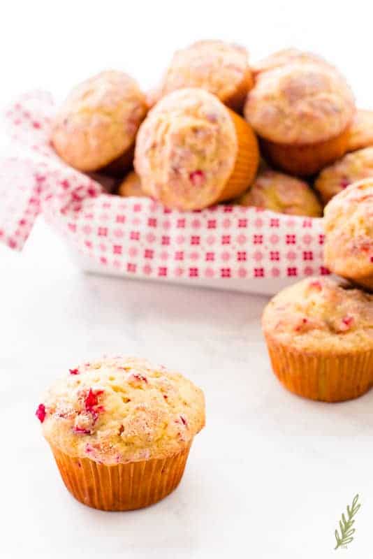 A Vanilla-Cranberry Muffin sits by itself in front of a basket filled with more muffins