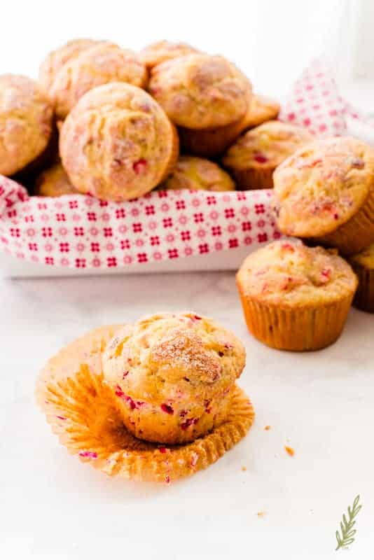 The wrapper of a Vanilla-Cranberry Muffin is pulled back to reveal itself. A basket of more muffins is in the background
