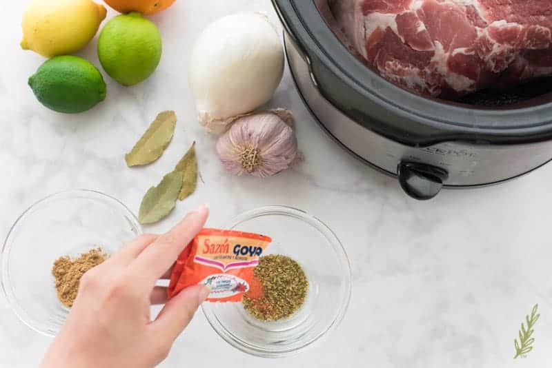 A spice blend is made in a glass bowl. A slow cooker has the the pork which will be pulled after cooking. Citrus, aromatics, and bay leaves are next to the slow cooker. 