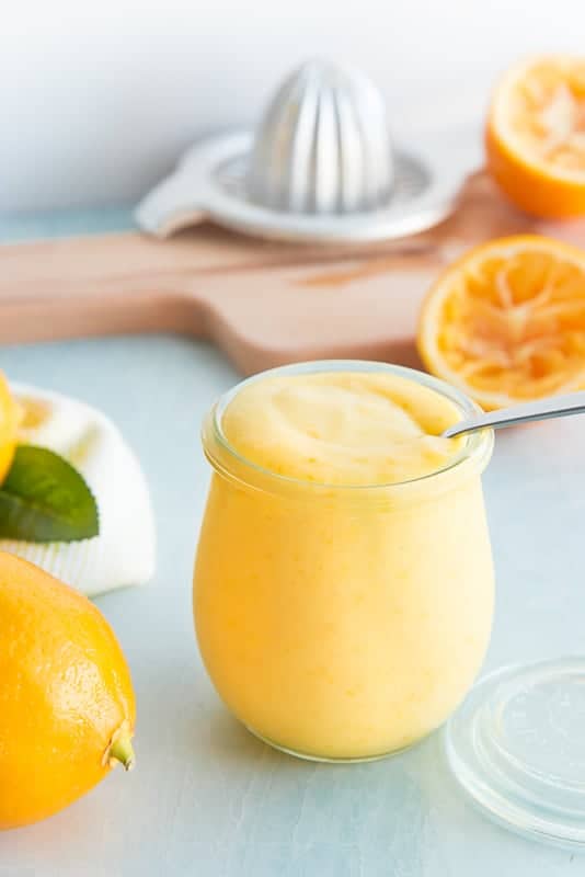 A spoon inserted into a clear glass jar of Meyer Lemon Curd surrounded by juiced lemon halves.