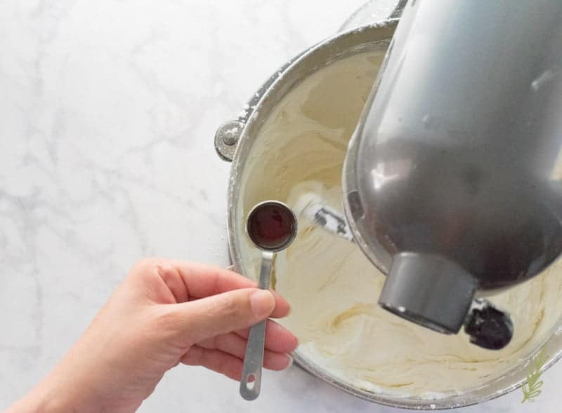A hand holds a teaspoonful of vanilla extract which is to be added to the butter-sugar mixture in the bowl.