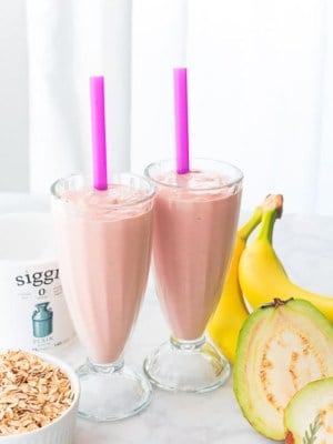 Two glasses of Guava Banana Oatmeal Breakfast Shake are surrounded by their ingredients.