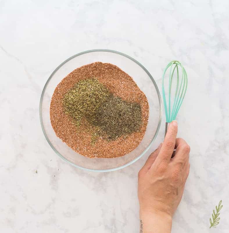 Dried herbs are added to a glass bowl with the rest of the Meat Spice Blend