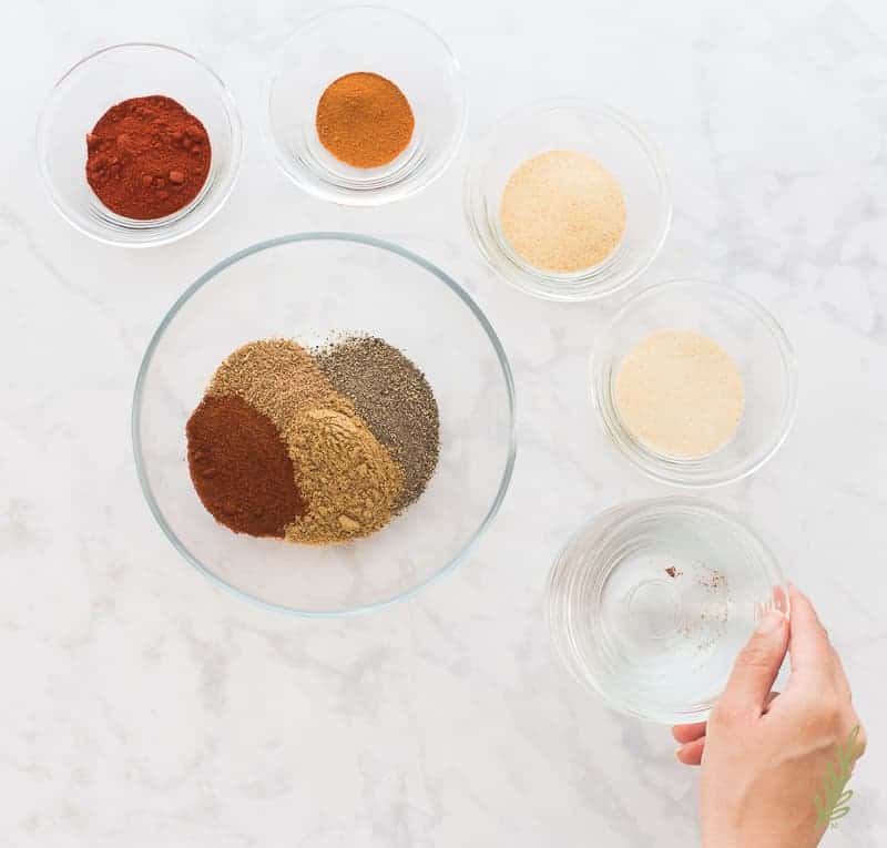 Red chili powder is added to a bowl of multi-colored spices