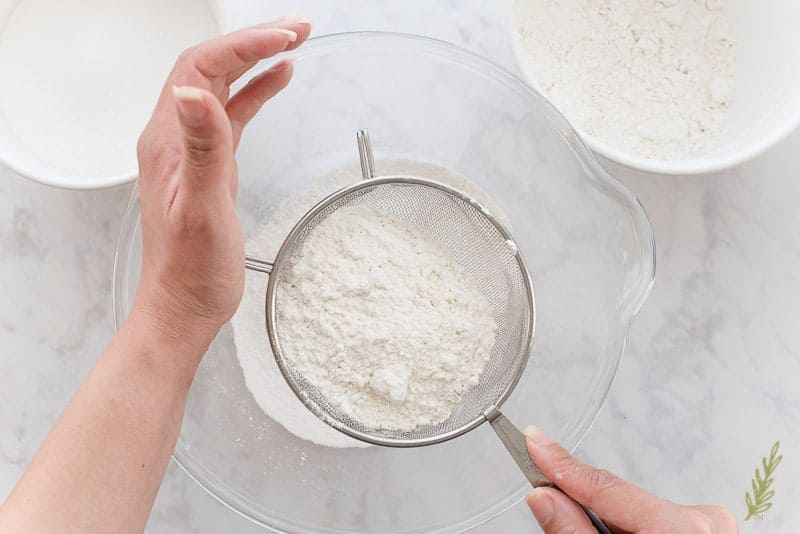 A hand holds a sifter filled with the cake's dry ingredients over a clear glass bowl.