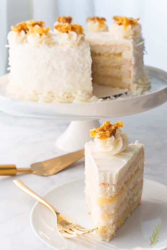 A slice of Piña Colada Cake has a bite removed. It's on a white plate which has a gold fork propped on it. The rest of the cake sits in the background