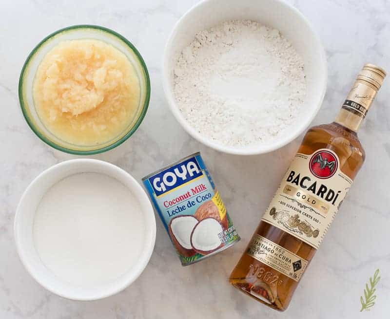 Piña Colada Cake ingredients from left to right: a white bowl of sugar, a glass bowl of crushed pineapple, a can of coconut milk, a white bowl with flour and baking powder, a bottle of gold rum