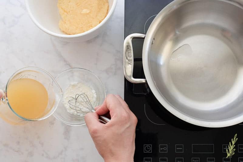 A portion of the pineapple juice-rum mix is added to a small glass bowl of cornstarch. A hand uses a whisk to mix the two together. To the right is a stainless pot on a burner. Above in a white bowl is the drained crushed pineapple.